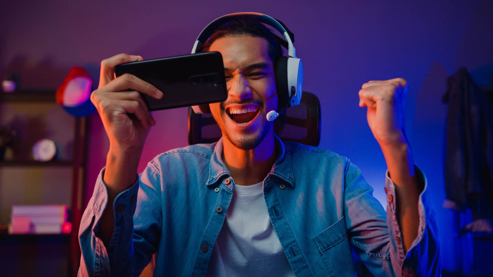 man in blue jacket and white shirt wearing headphones as he games and stares at phone while cheering