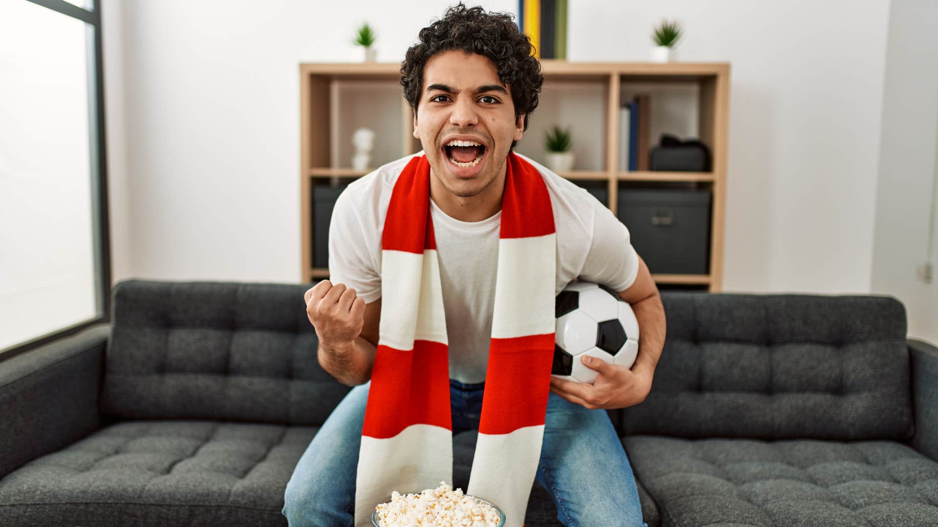 man in white shirt jumping as he watches soccer game and holds soccer ball with scarf around his neck