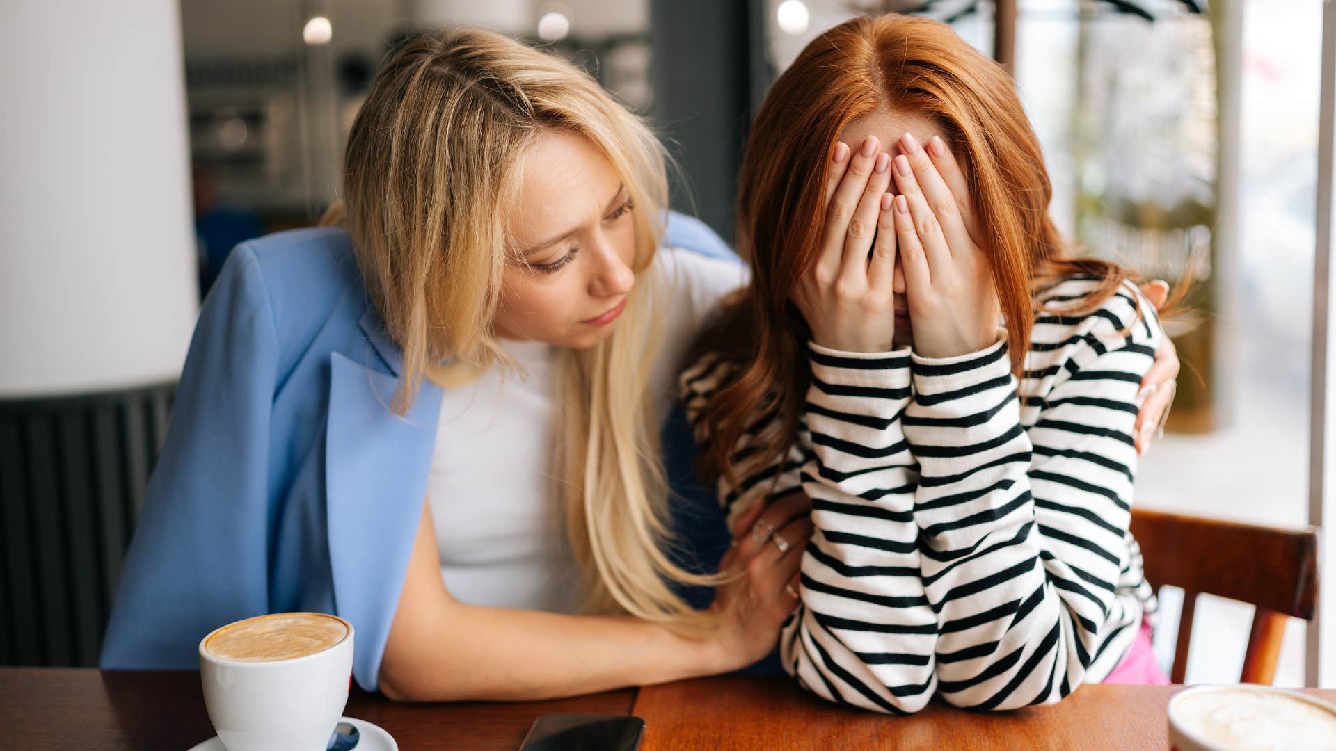 woman in black and white stripped shirt having a victim mentality as she cries and friend in blue jacket comforts her in a cafe