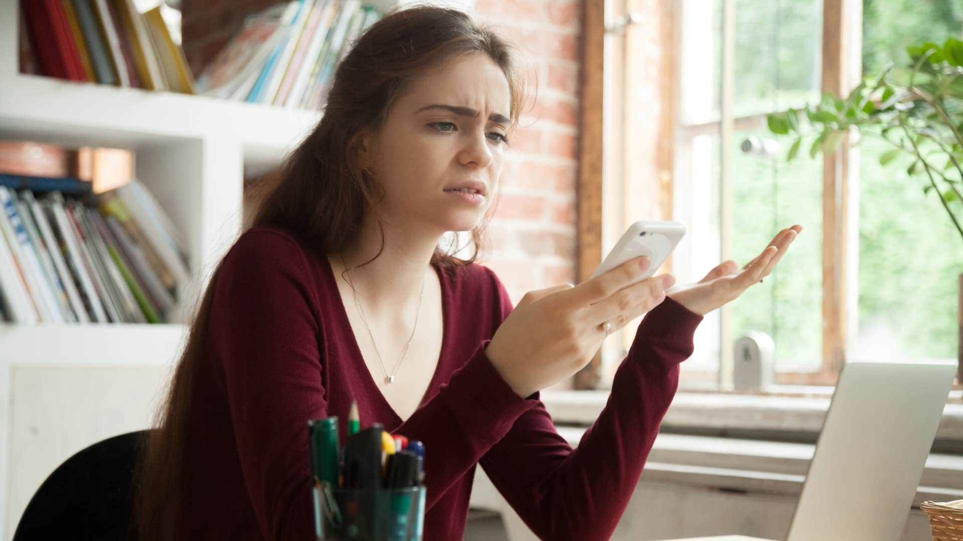 woman in red shirt looking annoyed at her phone as her friend uploads the worst photos of her