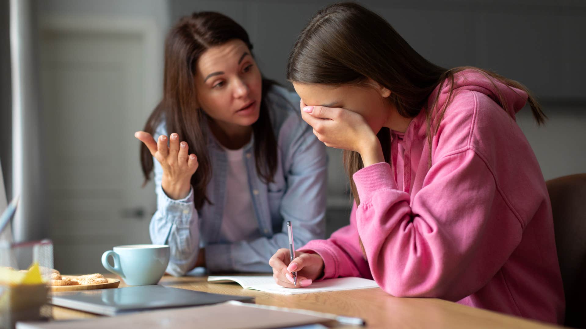 mom in blue cardigan telling daughter whose upset that she's too sensitive as daughter hides her face and writes
