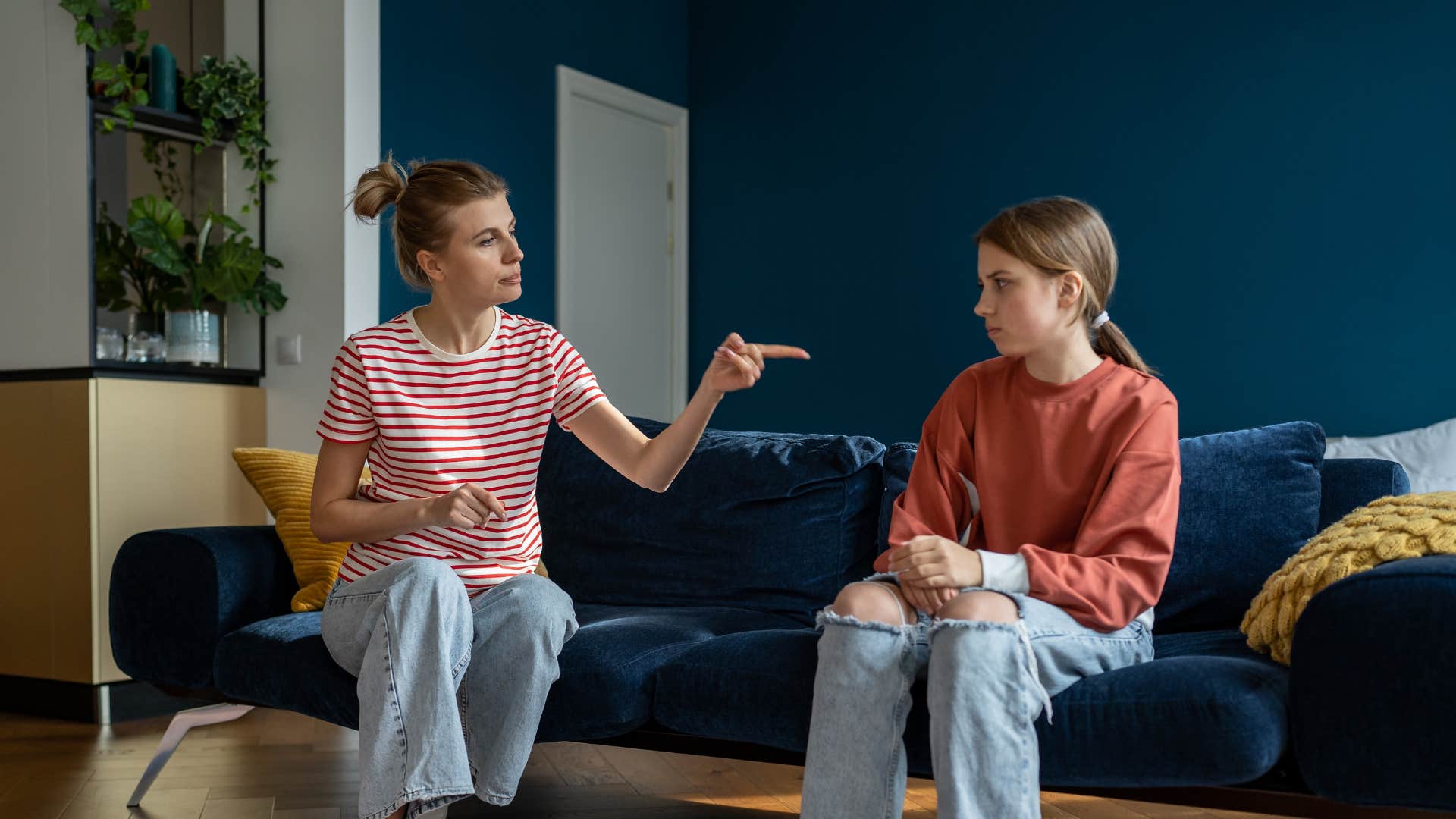mom in stripped shirt scolding daughter in red shirt as she sits on the couch saying whatever happens here stays here