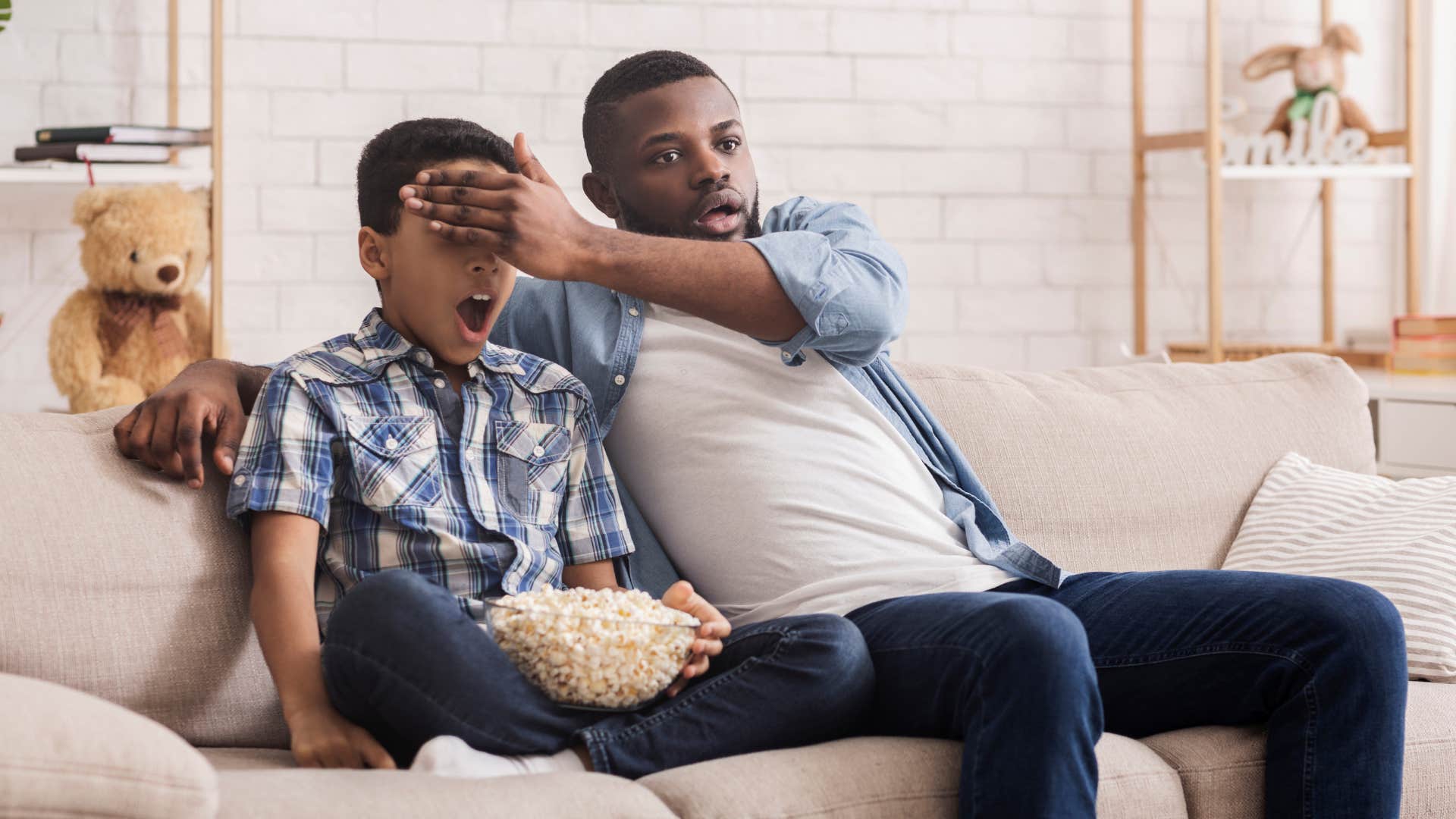a dad watches television with his son and covers his eyes over an inappropriate scene