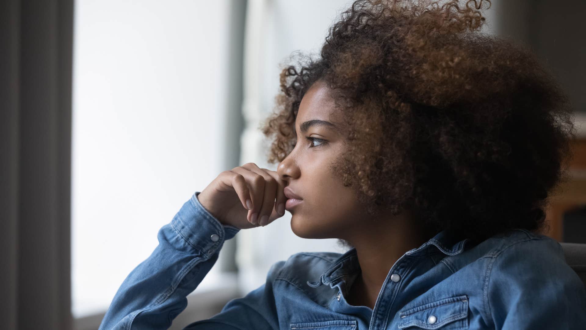 woman with curly hair in denim shirt feeling disconnected from others as she stares out the window thinking