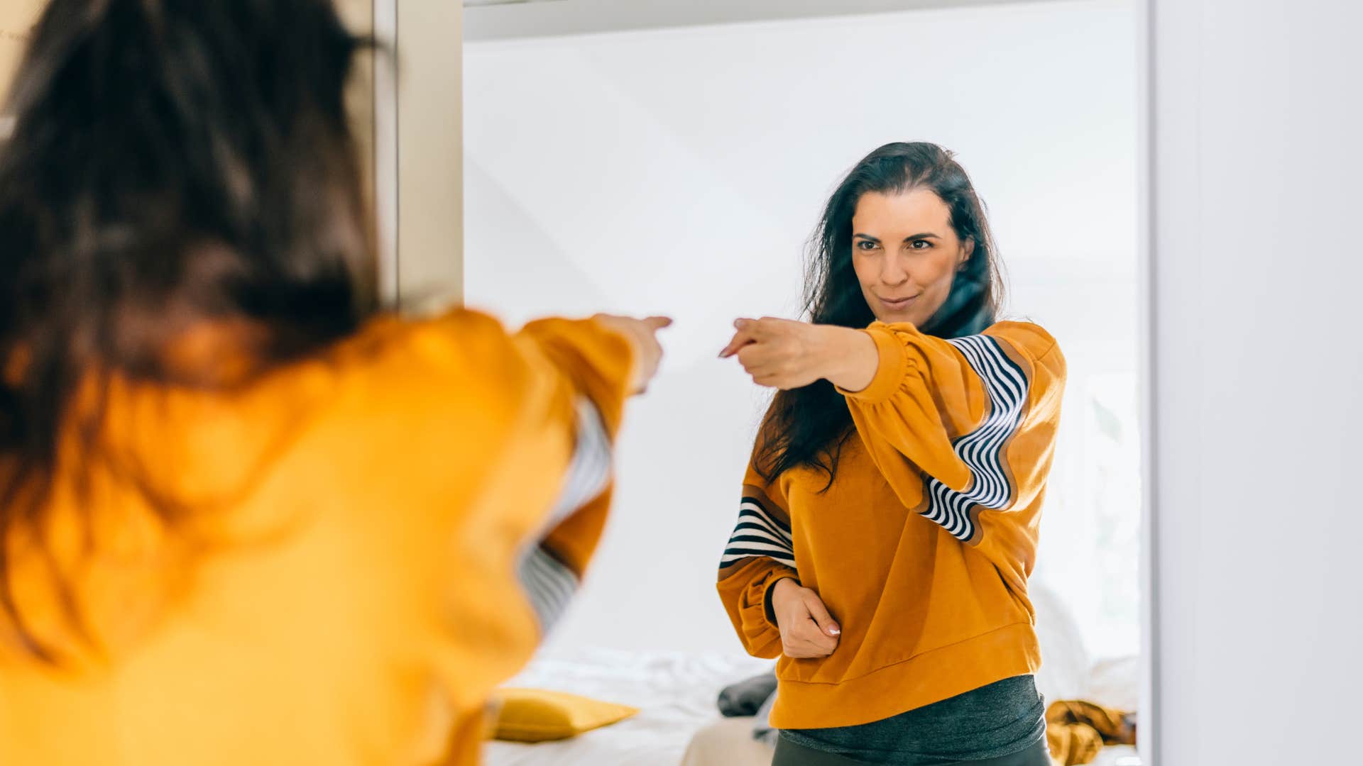 woman in orange shirt pointing to the mirror as she smiles and expresses how proud she is of herself