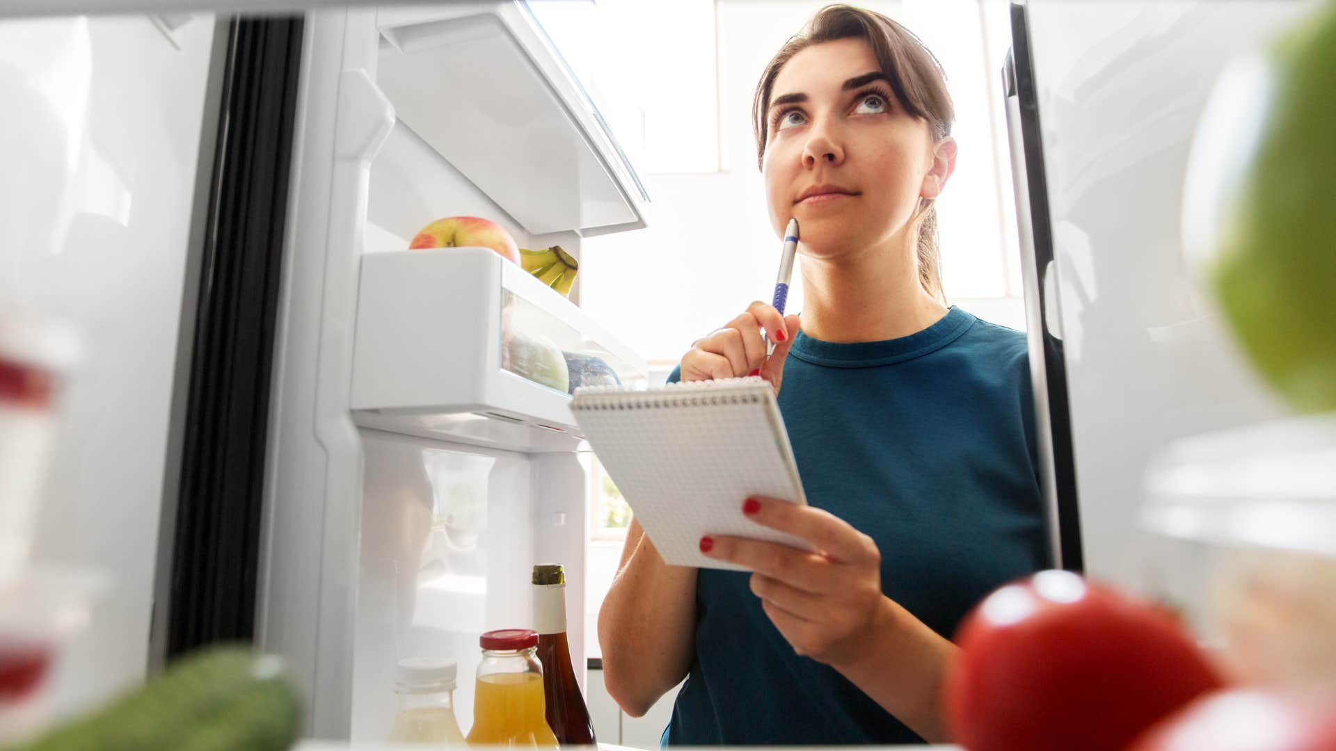 woman writing a list while looking in her fridge