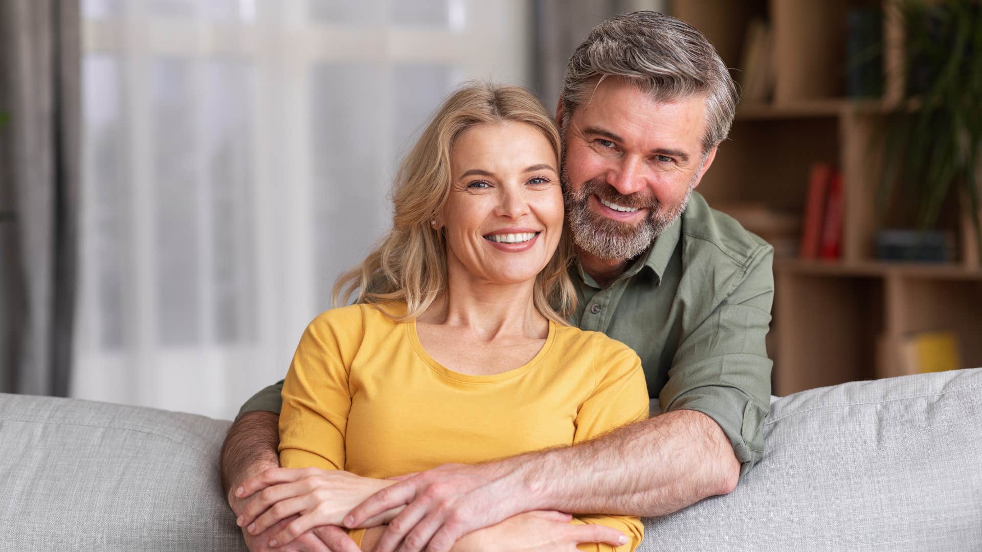 man in green shirt being emotionally available to woman in yellow shirt as he hugs her from behind as she sits and smiles