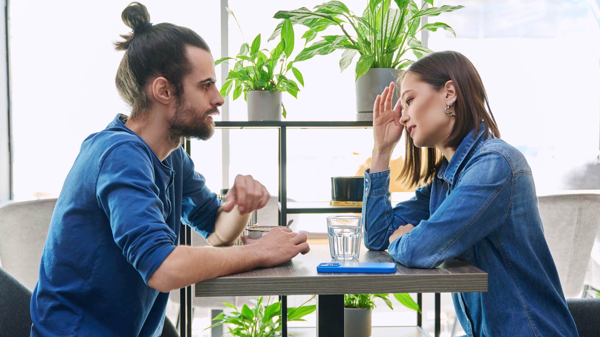 couple in jean shirts sitting across from one another looking tense as woman feels butterflies around him
