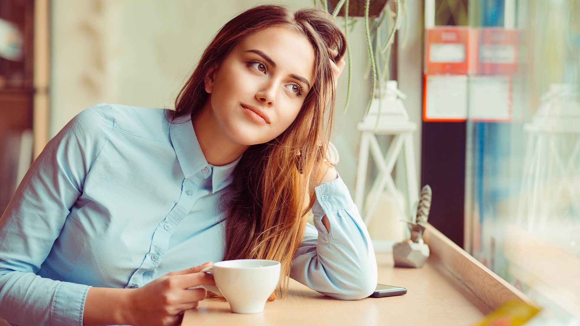 woman in blue shirt finding herself daydreaming more often than not as she looks out window with cup of tea in hand