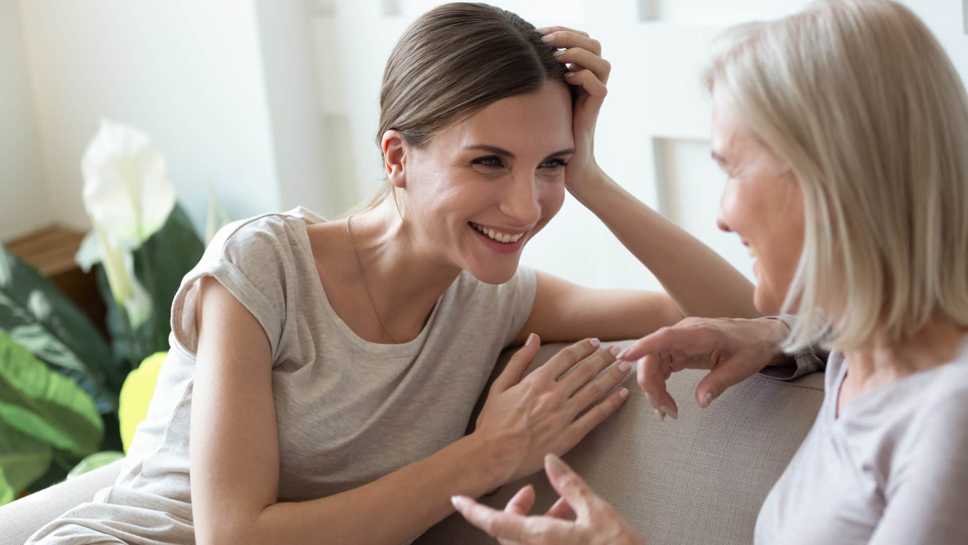older woman in gray shirt telling younger woman on couch god love him he can't help it as they gossip and laugh