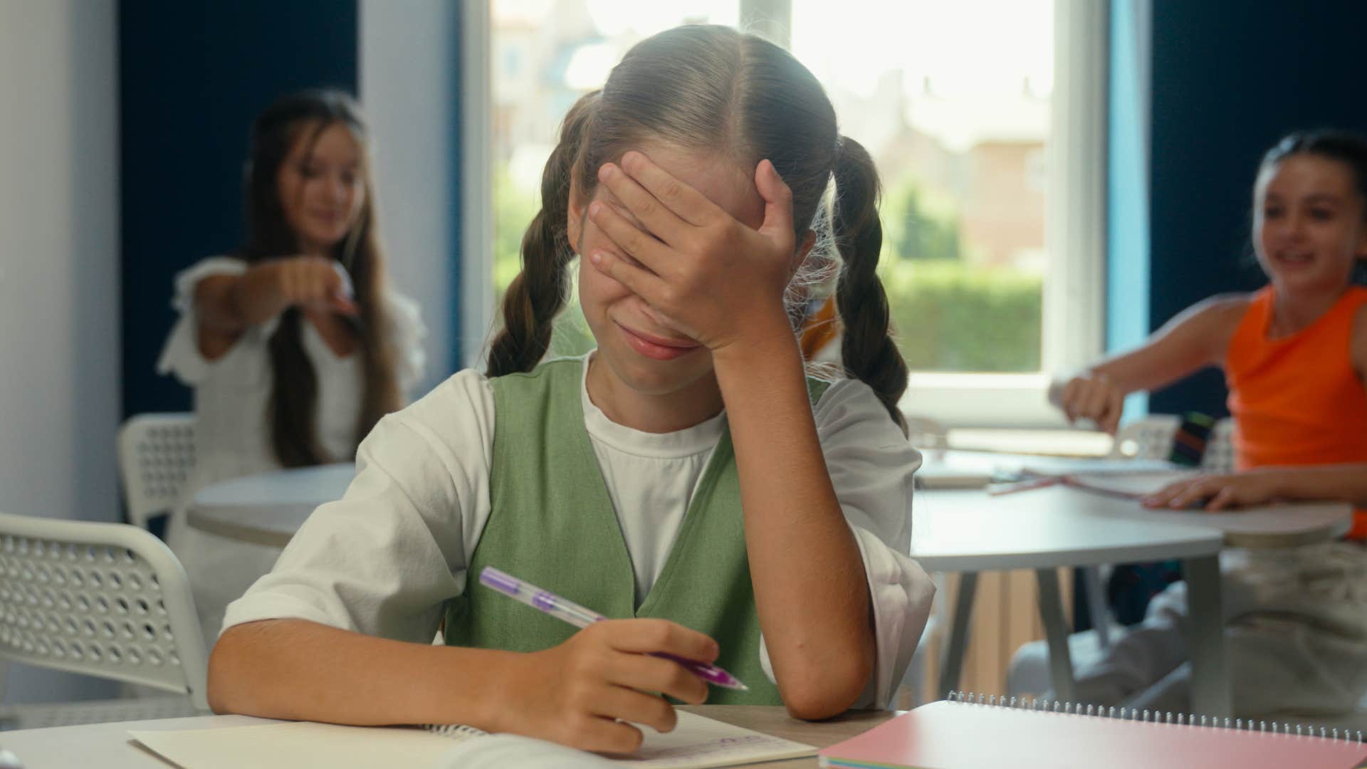 Upset girl holding her face in her hands around friends who are cruel to peers.