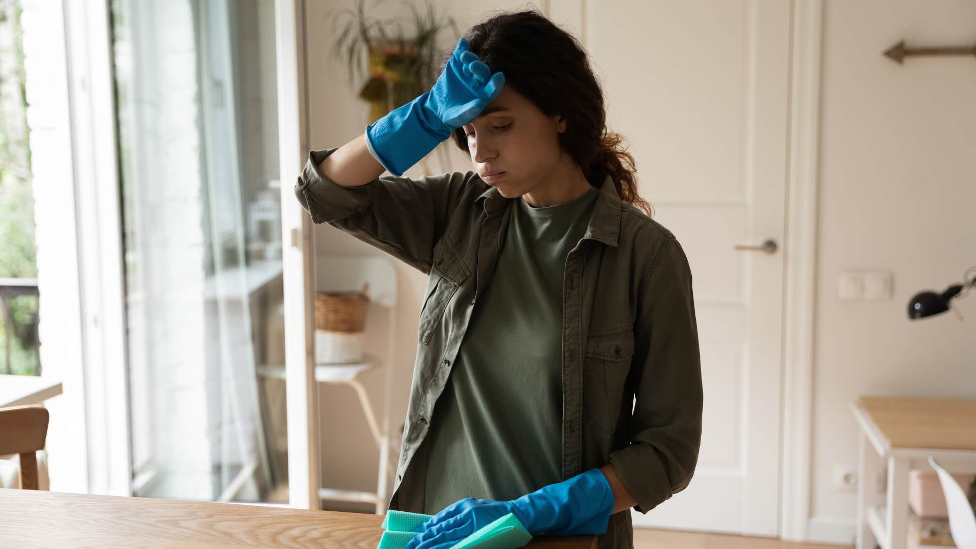 woman in green dark outfit and blue gloves keeps on taking on the role of a manager as she wipes forhead