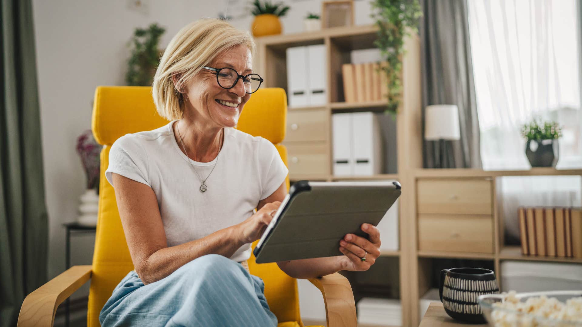 woman reading or writing on tablet as she smiles with glasses and sits in yellow chair