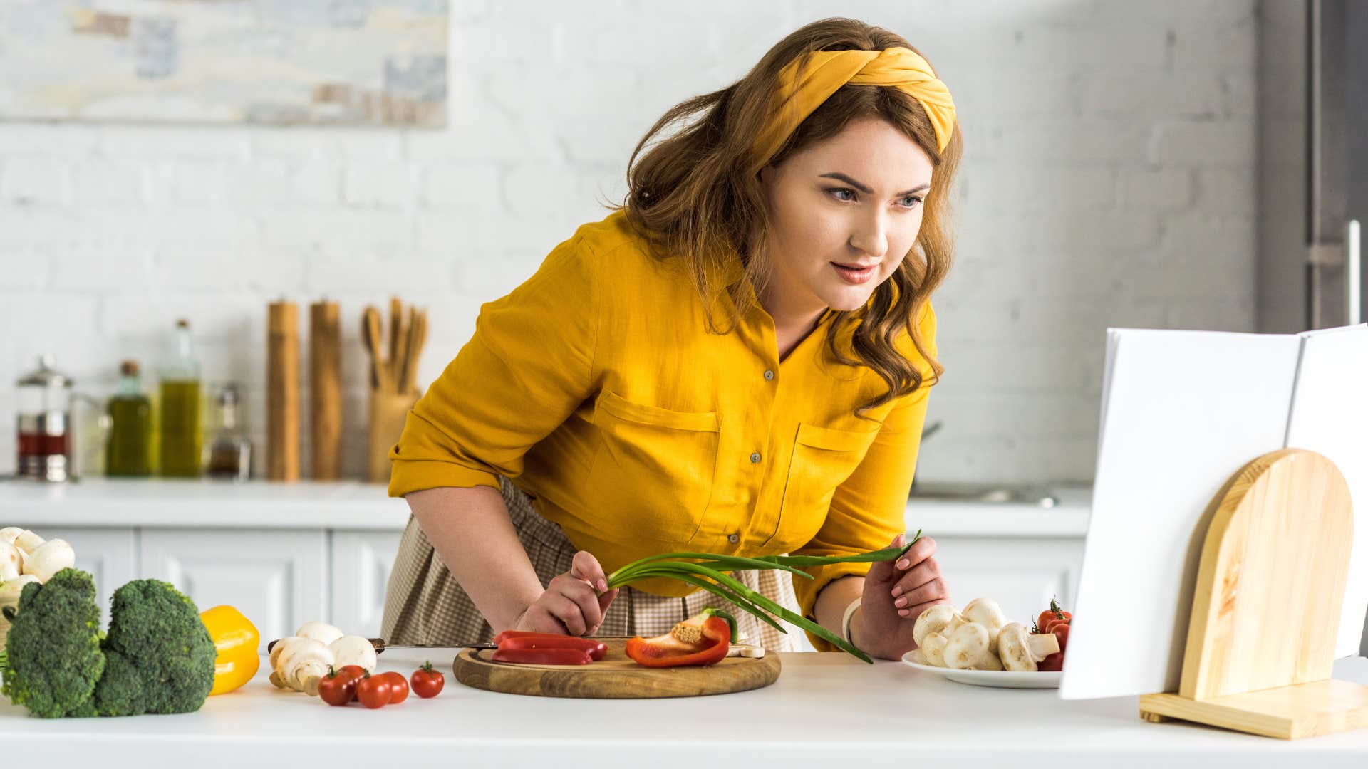 woman in yellow shirt having a collection of cookbooks as she reads one while cooking