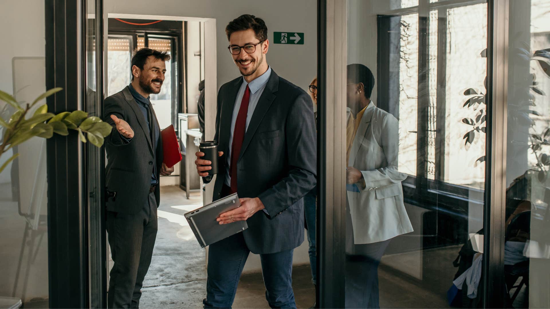 man in suit holding open door for others as man in suit and red tie walks in