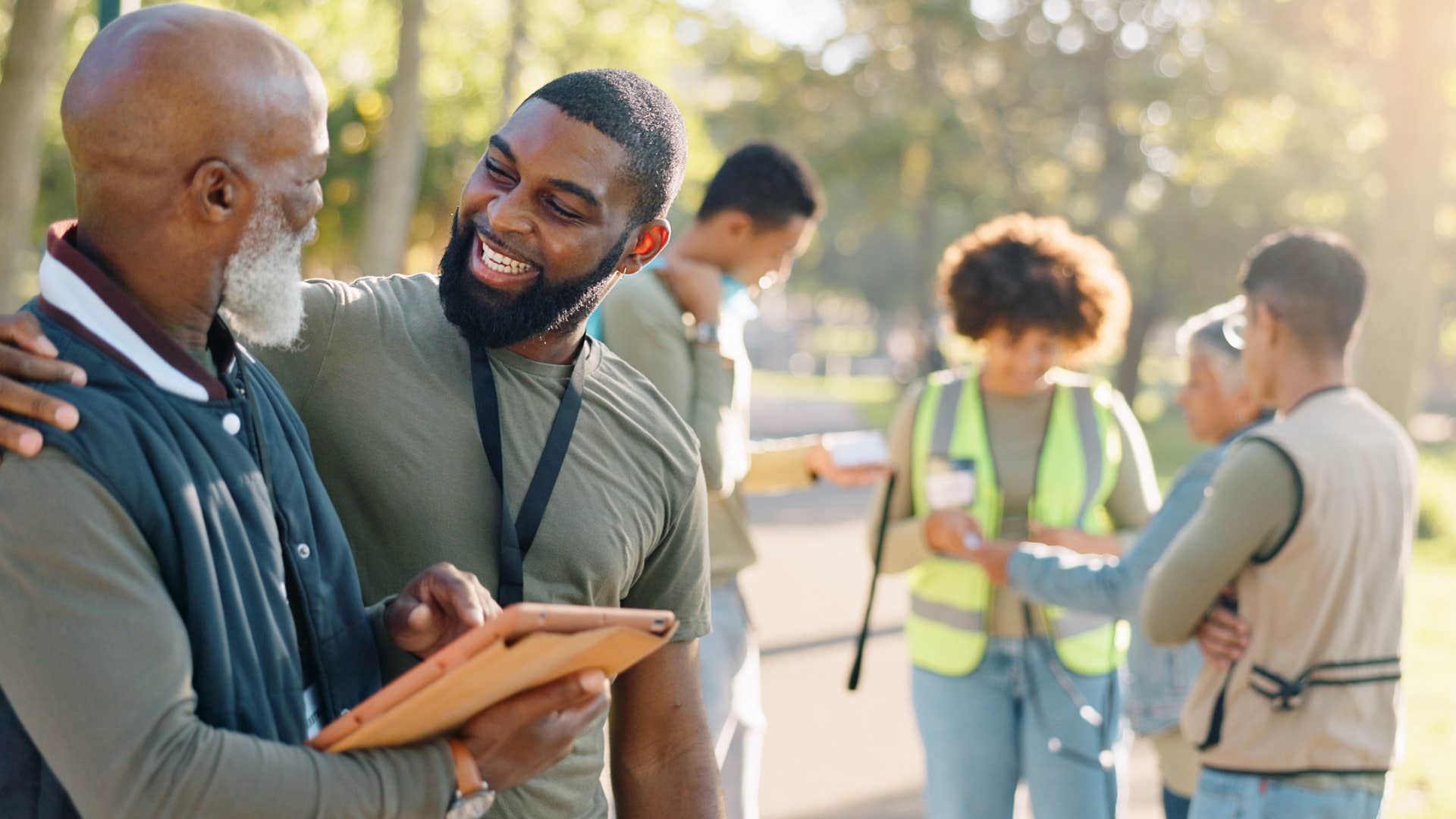 man in gray going out of his way to include others as he side hugs man with clipboard and smiles