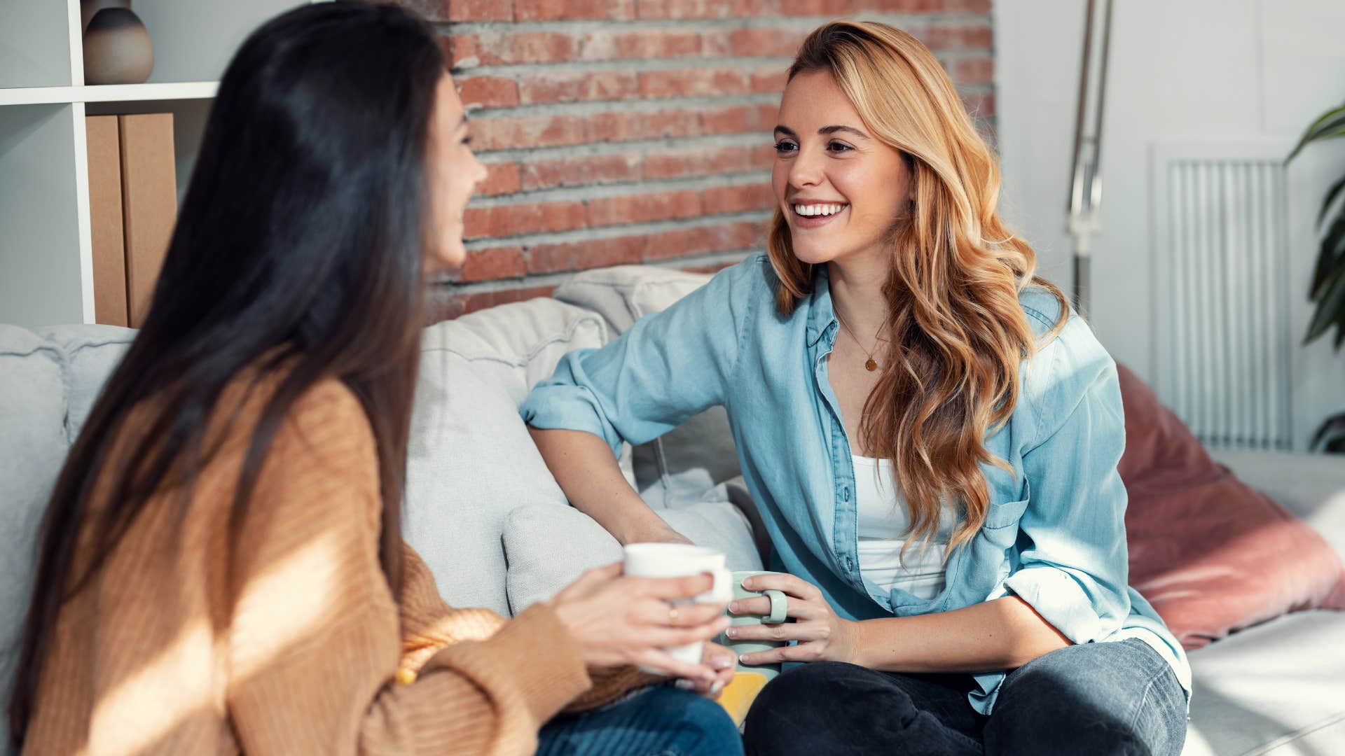 woman in light blue jacket speaking kindly about others behind their back with friend in brown shirt as they sit on couch and smile