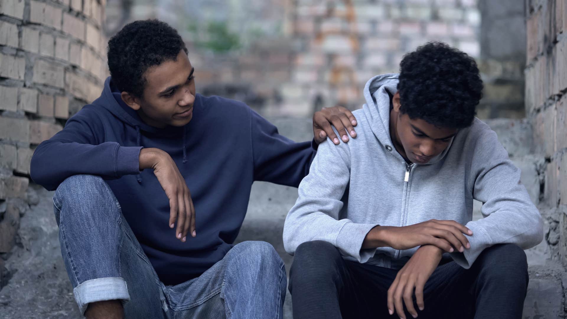 man in light hoodie saying i'll be okay while looking down as friend in blue hoodie comforts him