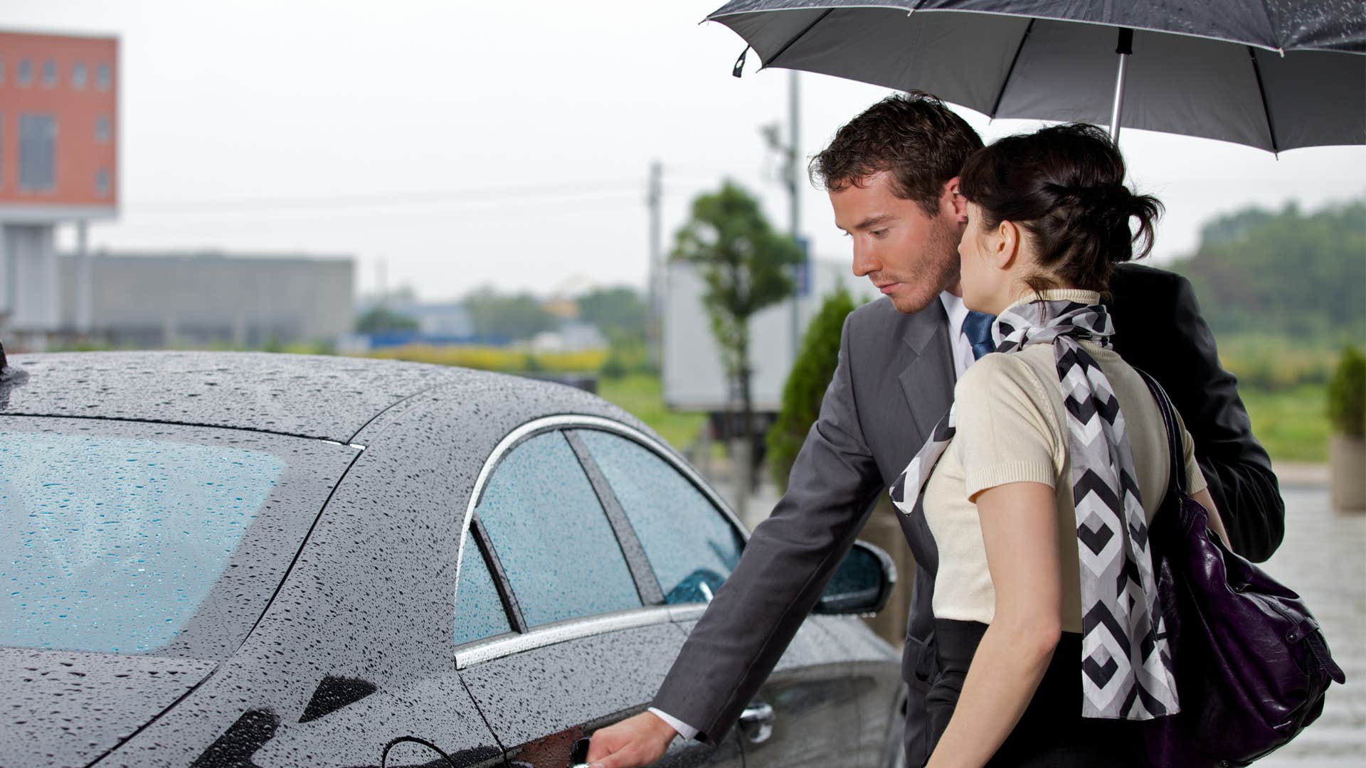a man uses the umbrella from his car to let his family member into his car while it is raining