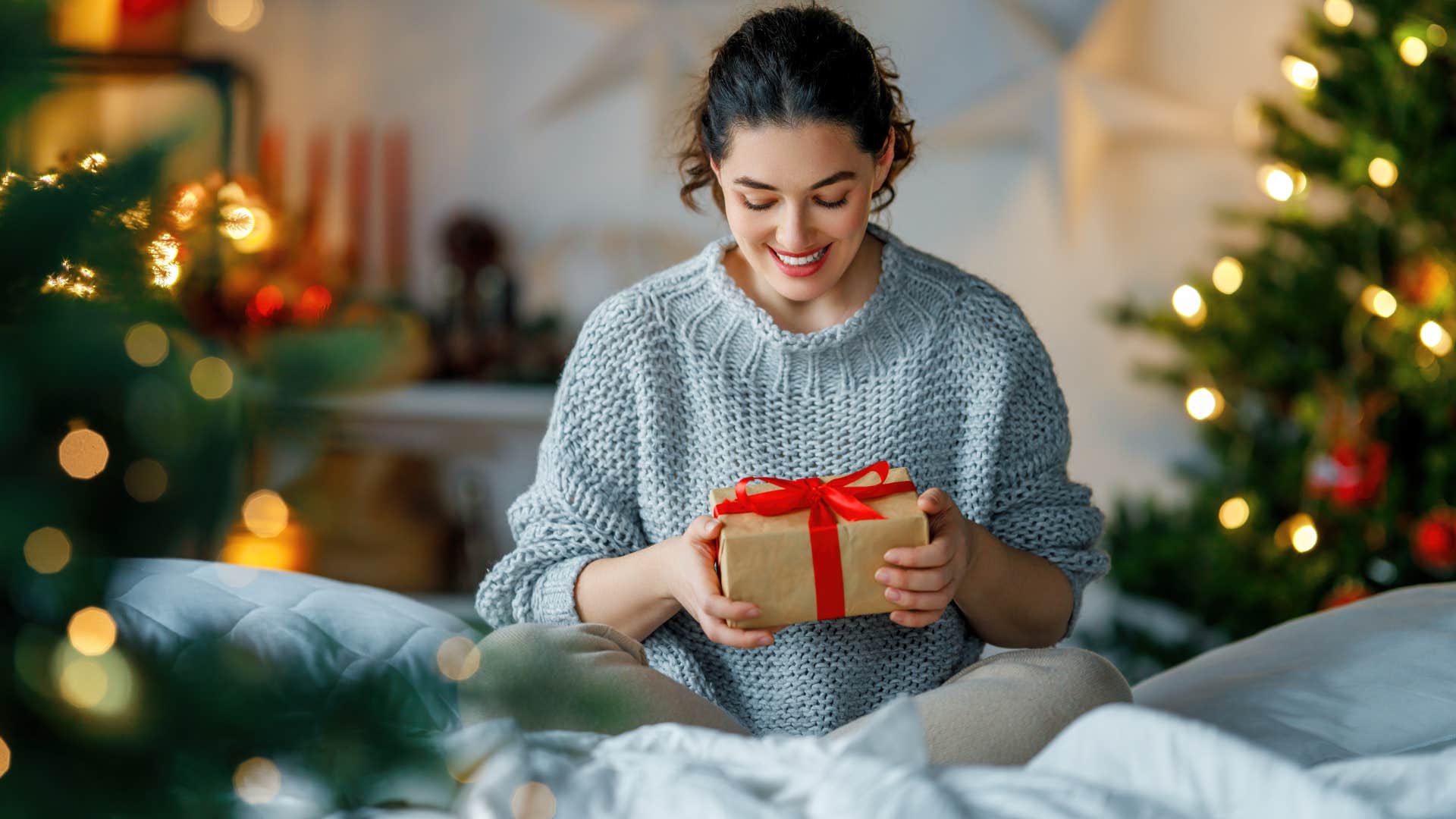 woman in gray sweater looking at fancy holiday gift as she smiles 