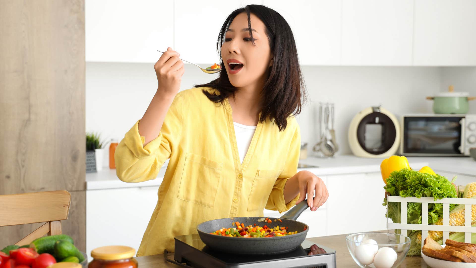 woman in yellow shirt cooking with one pan as she eats from spoon