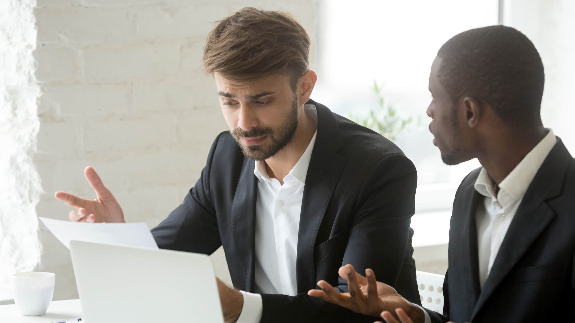 man in suit having to talk as he talks to coworker clearly annoyed