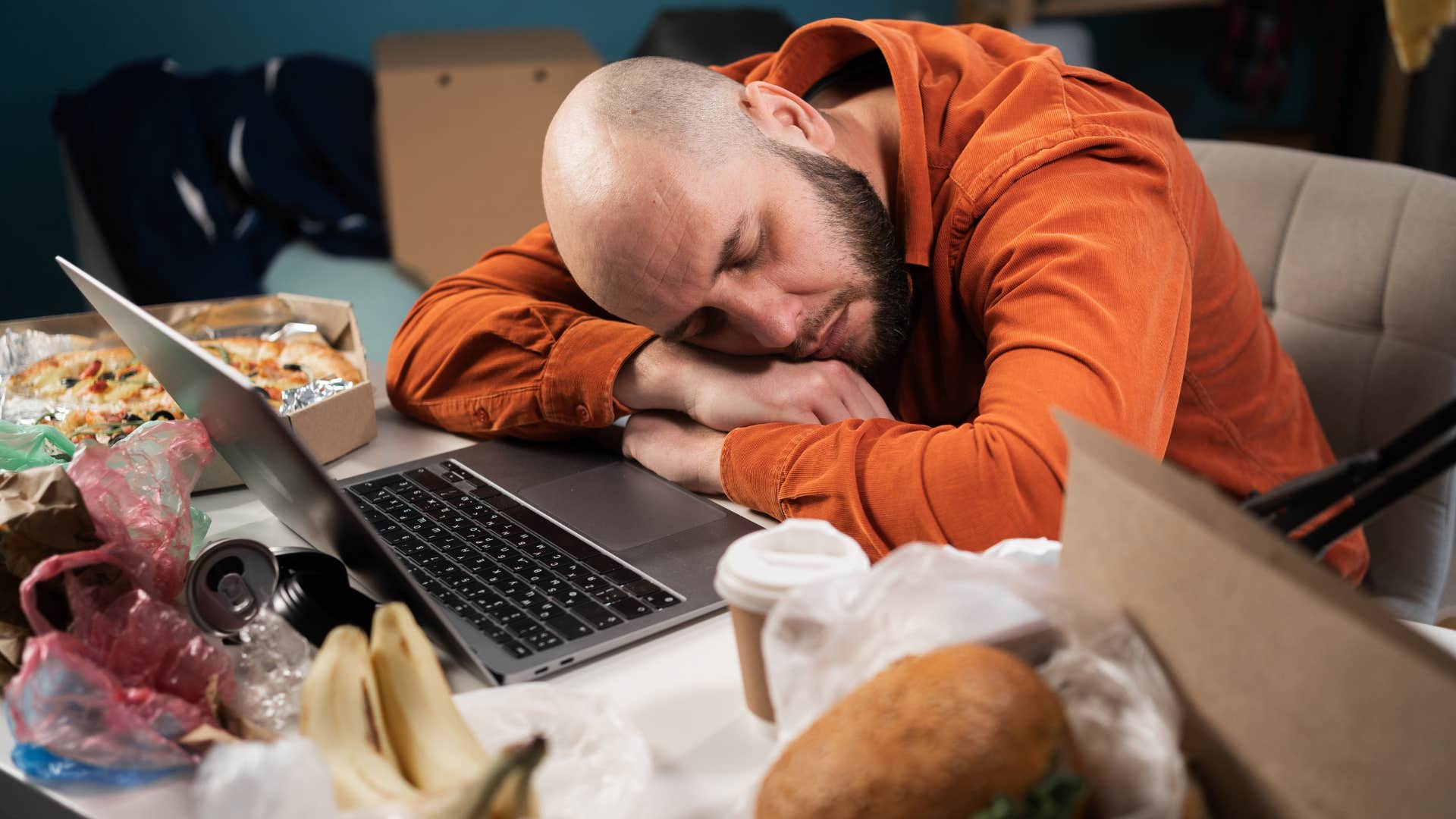Man sleeping at his laptop surrounded by clutter.