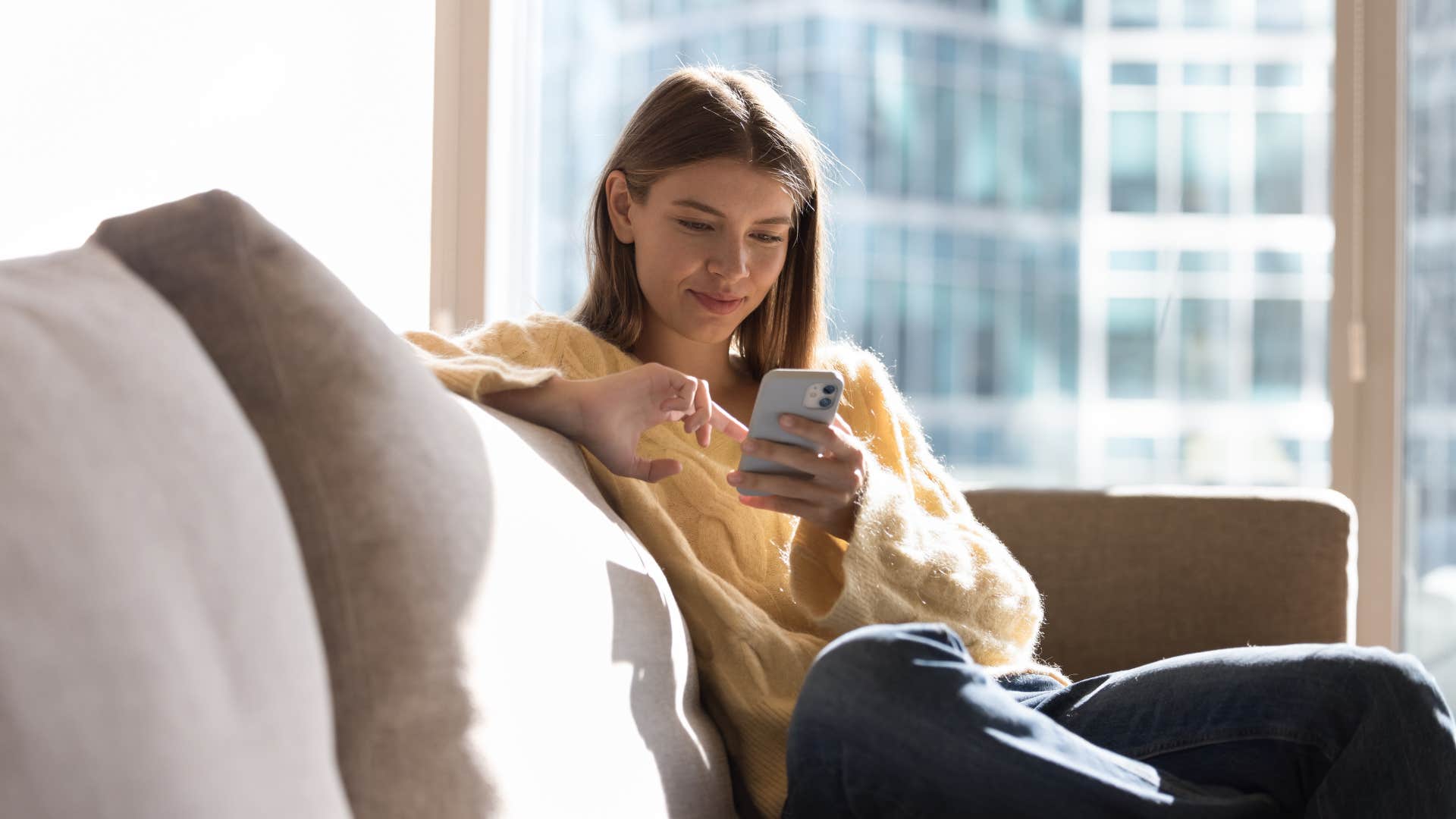Woman sitting on her couch next to large windows.
