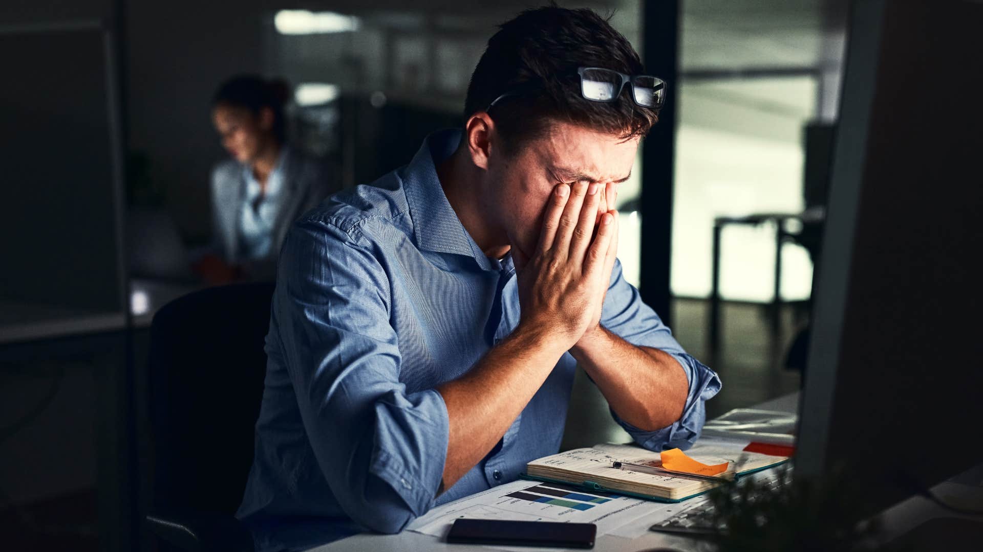 man in blue shirt staying at work longer than necessary as he rubs eyes