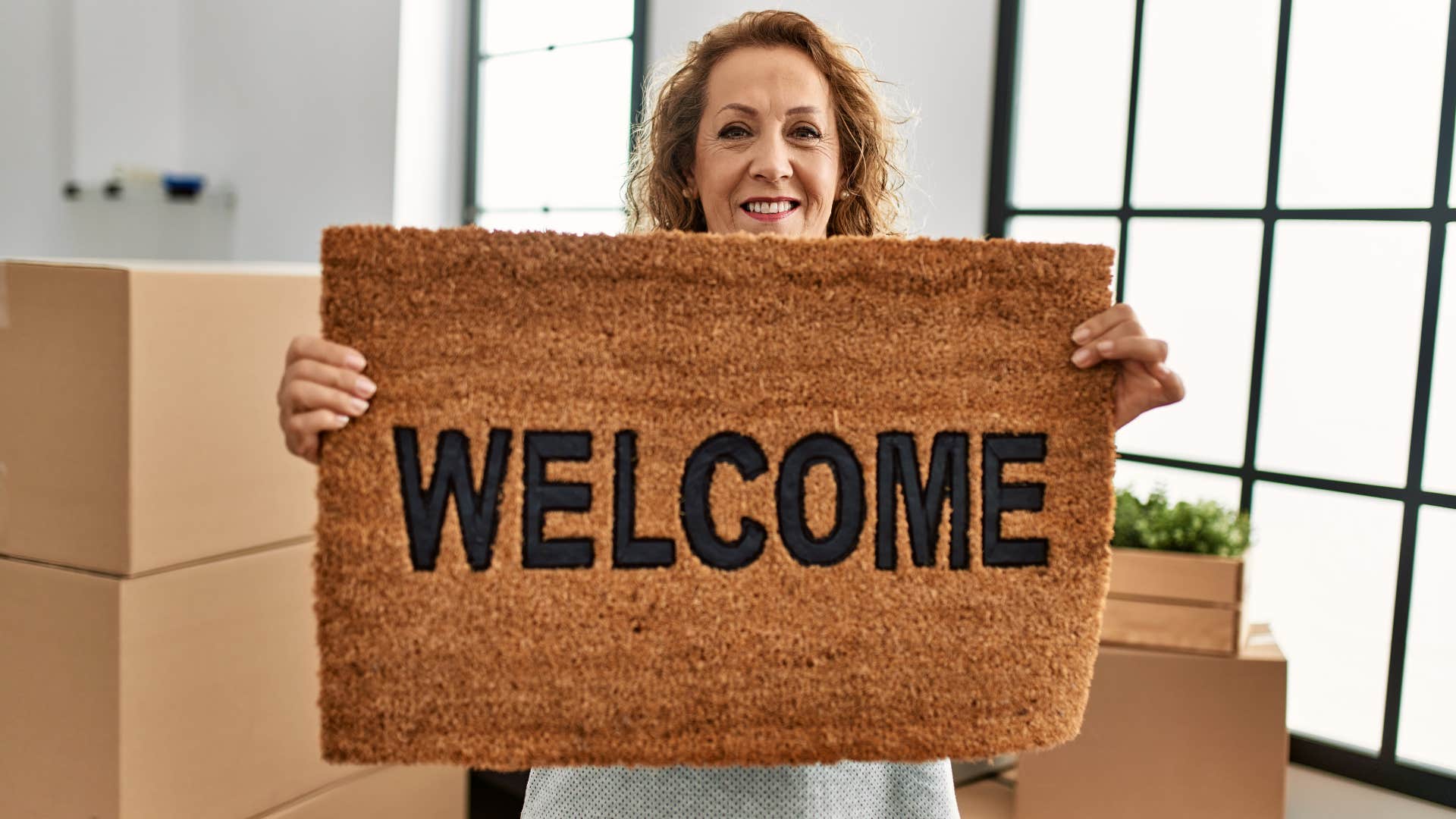 Woman smiling and holding up a welcome mat at home.