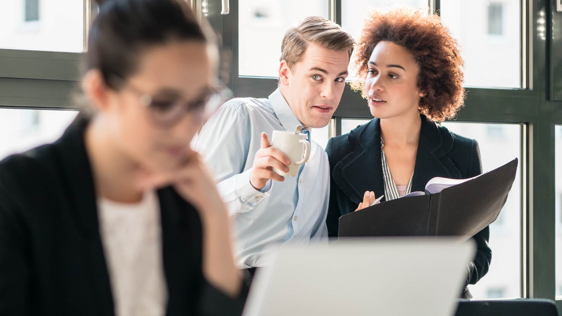 Co-workers gossiping behind a woman in an office.