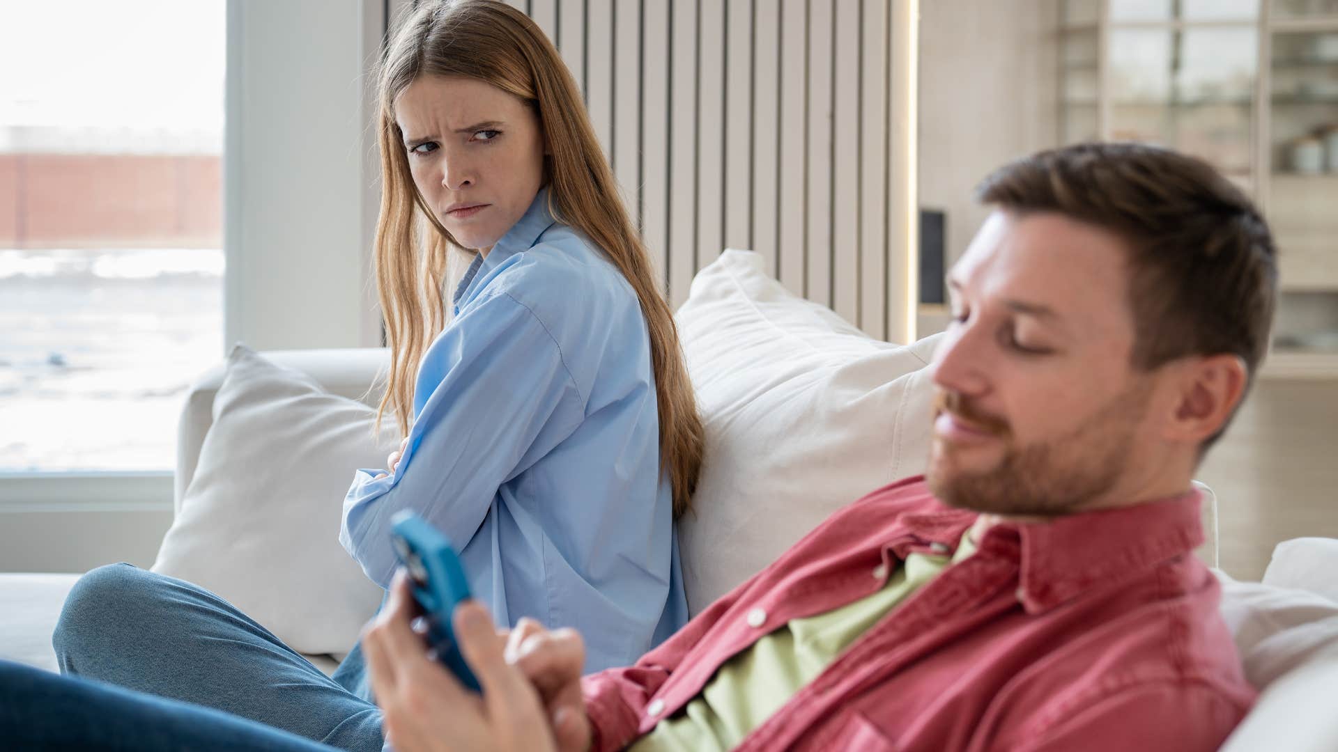 Woman looking at her husband who avoids arguments and conflicts on his phone.
