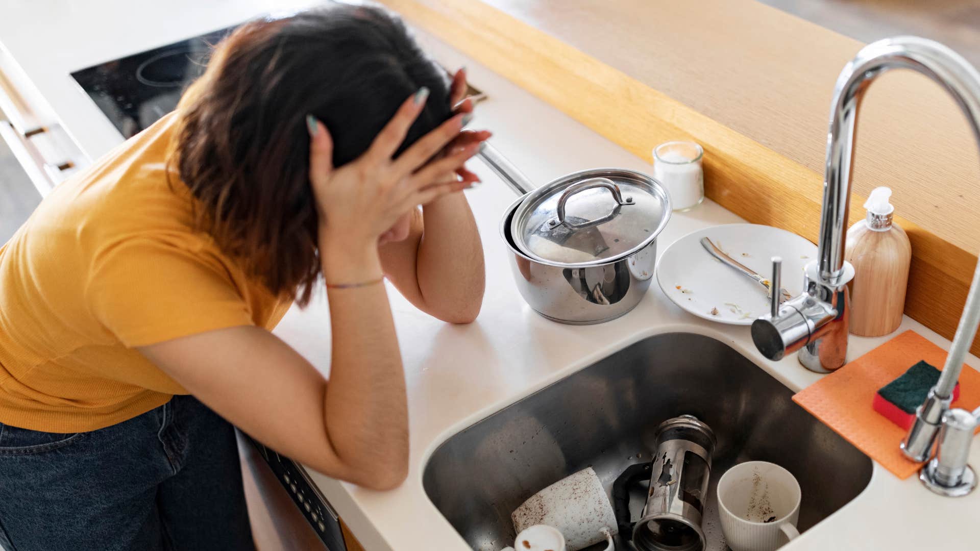 stressed woman holding her head around overflowing dishes in the sink