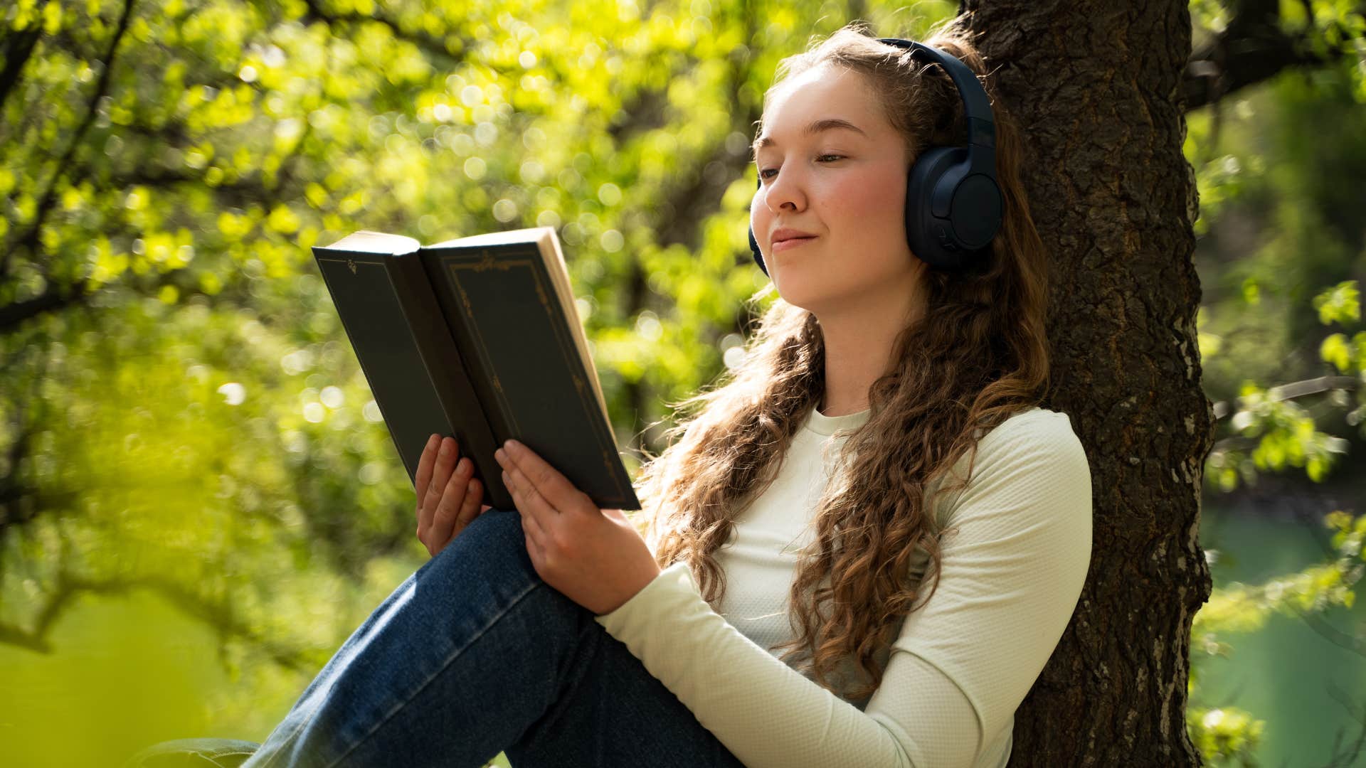 curious woman reading book against tree wearing headphones