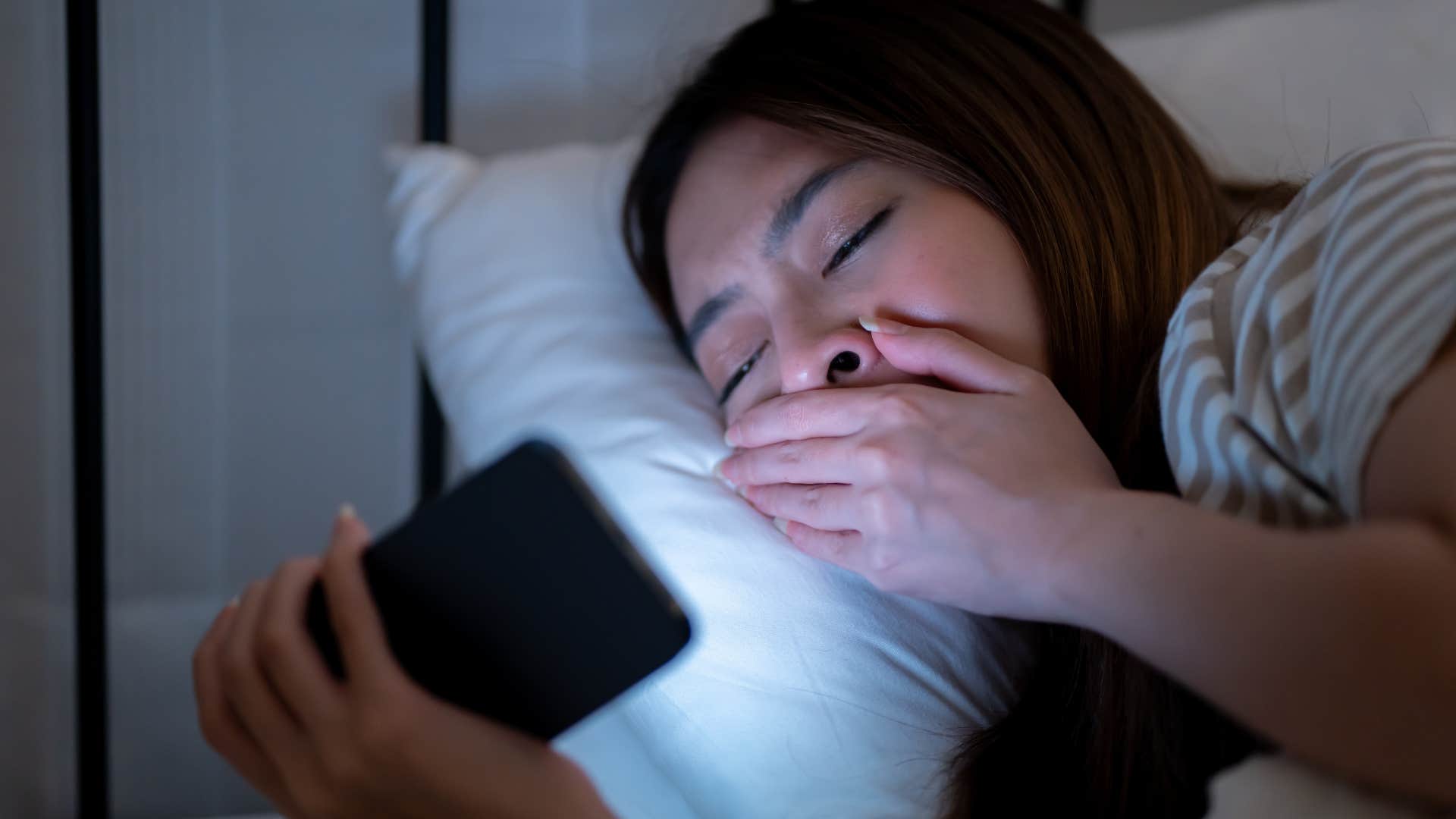 Woman whose sleep schedule is inconsistent yawning on her phone.