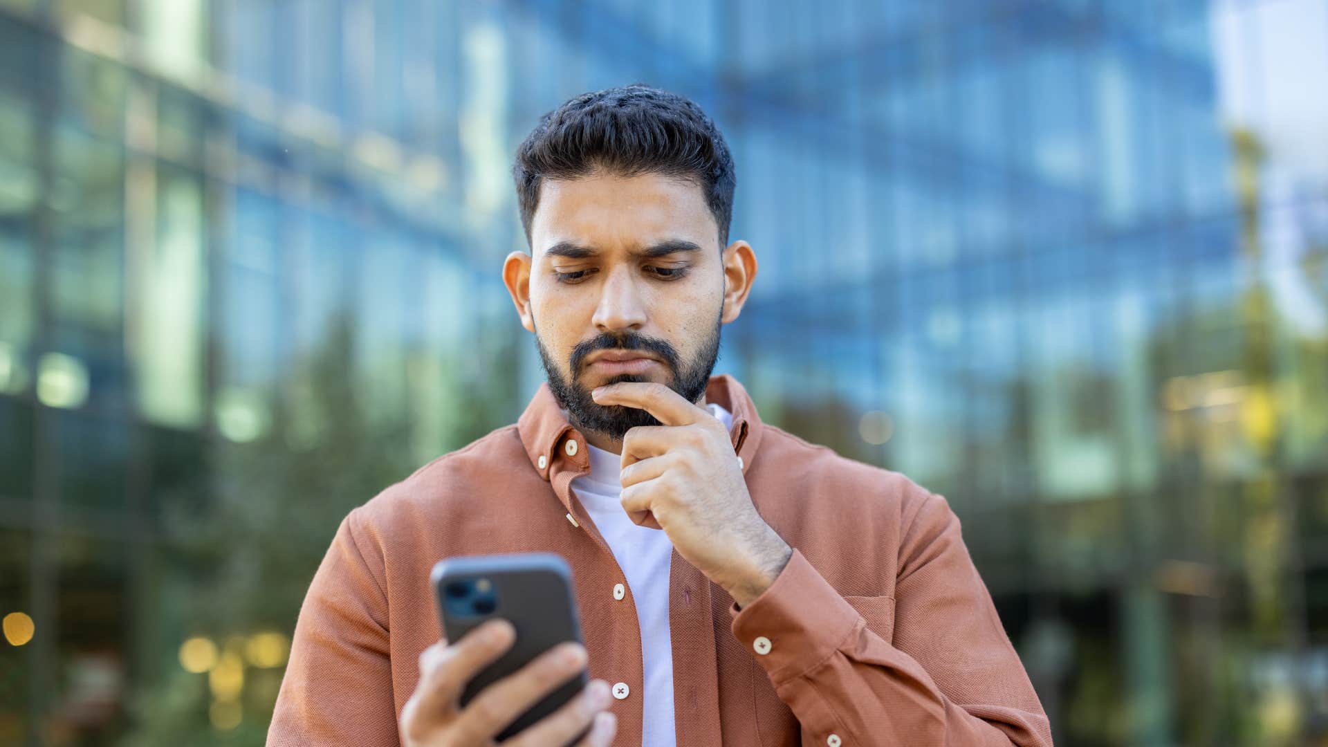 man in white shirt and pink-brown jacket looking at phone thinking of ways to make her day better
