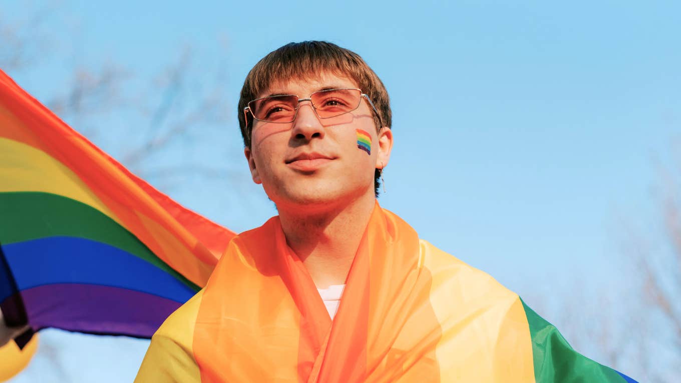 Mom Of A Boy Bullied For Being Gay Shares TK Ways All Parents Can Help Stop Anti-LGBTQ Bullying Teen boy wearing an LGBTQ pride flag wrapped around his shoulders, representing efforts to stop anti-LGBTQ bullying