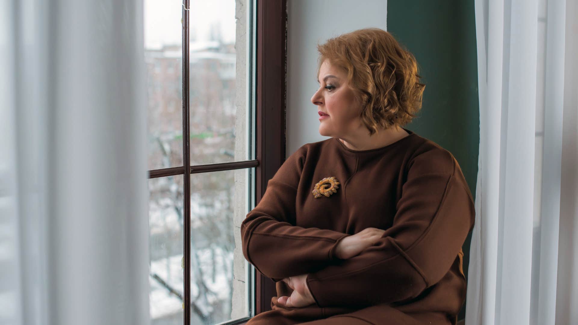 woman wearing an old brown sweater as she holds onto sentimental items out of guilt and stares out the window