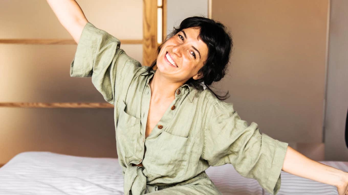 Happy young woman sitting on bed, smiling and spreading her arms in a bright minimalist room, illustrating the joyful habits of naturally minimalist people and intentional simple living.