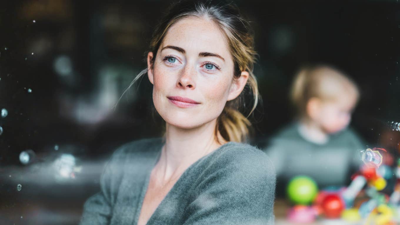 A calm woman gazes out the window while her child plays behind her, symbolizing self-care, emotional boundaries, and the power of learning to put herself first without guilt.