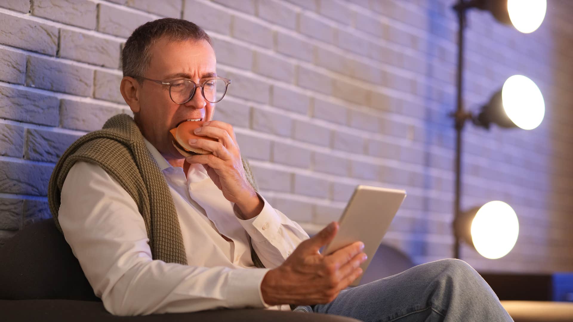 man in white shirt looking at tablet and eating hamburger as he doesn't cook real meals anymore