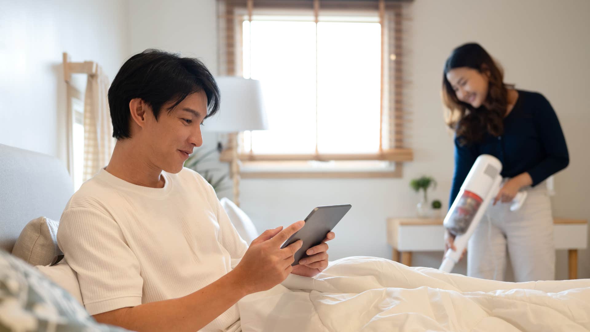 man on tablet in bed while wife vacuums floor