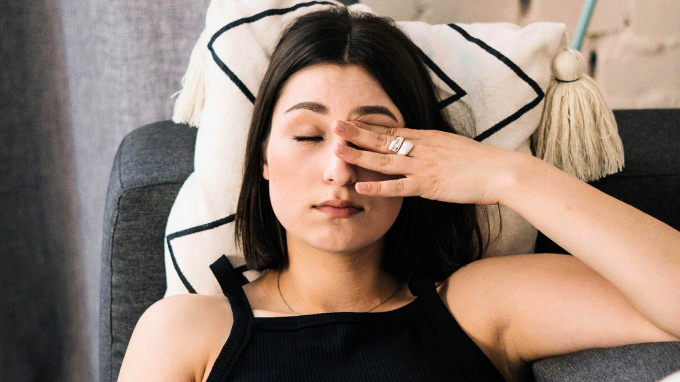 Exhausted woman rubbing her eyes on the couch, illustrating the quiet guilt people feel for resting due to habits learned in childhood.