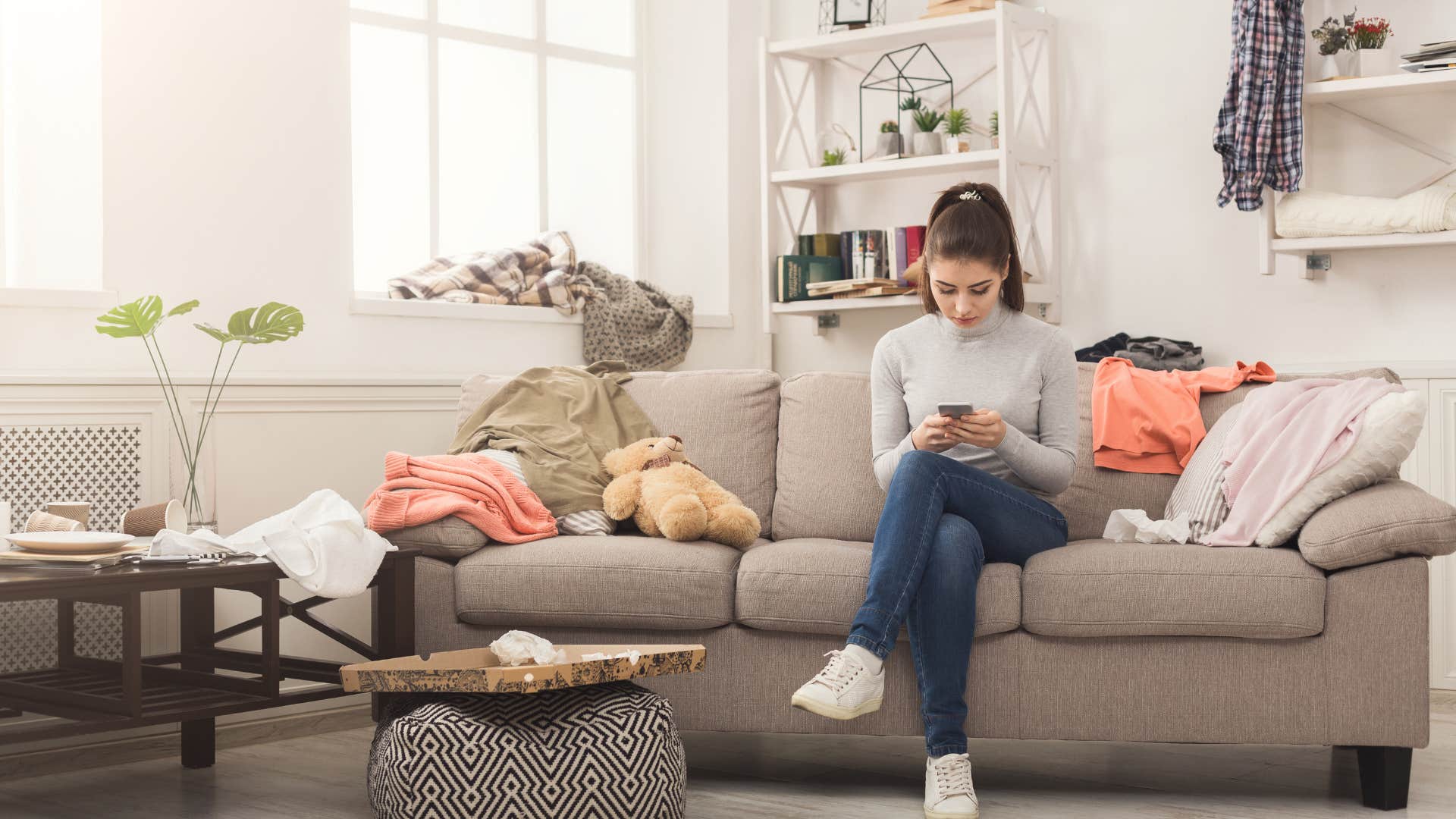 woman sitting in messy room on her phone
