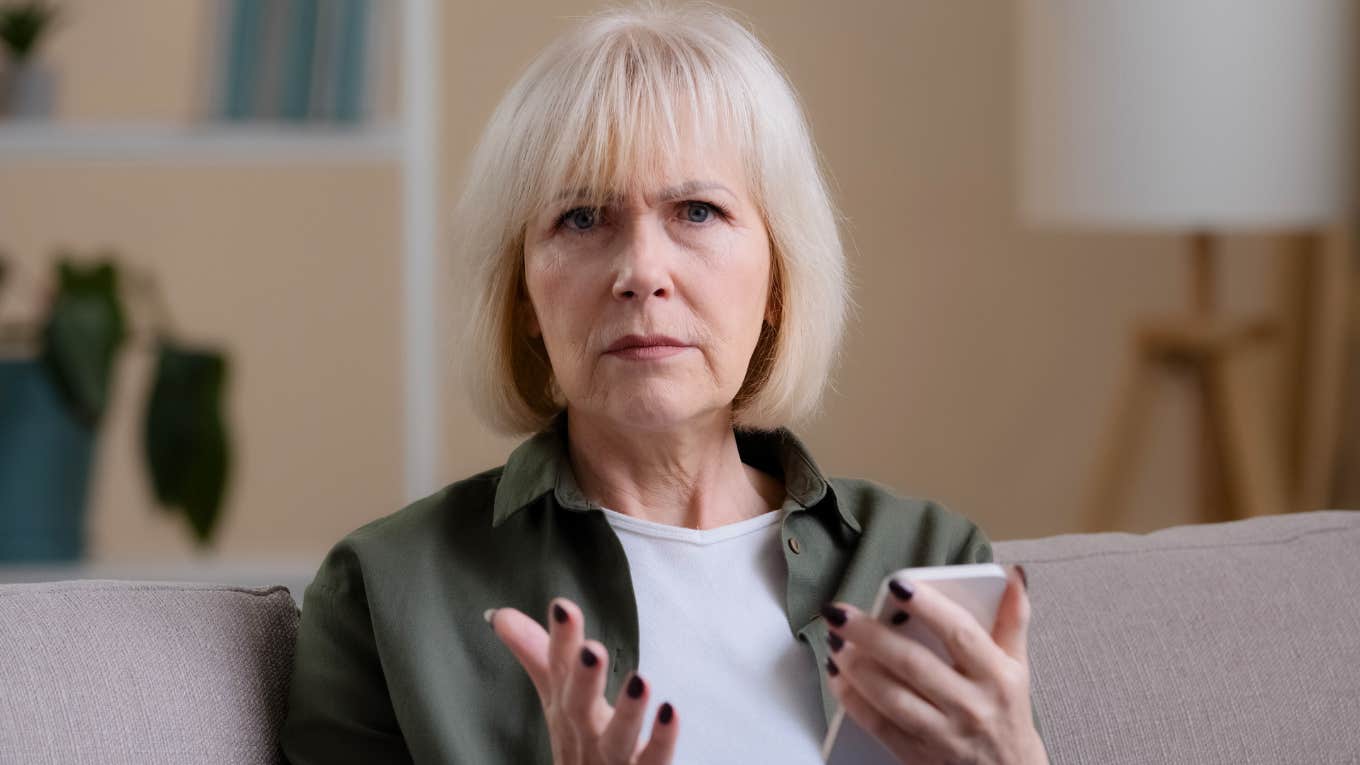 Serious older woman holding a phone on a couch becoming less patient as she ages