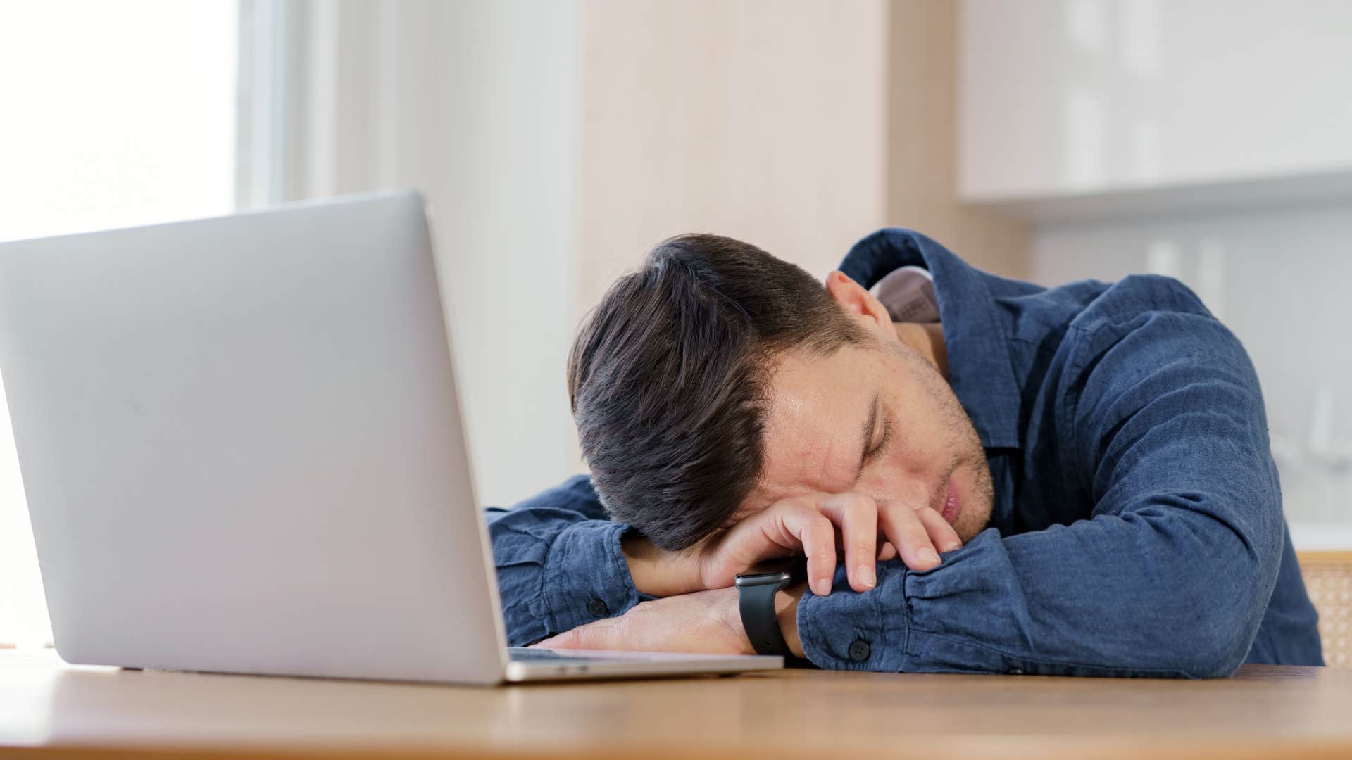 man in dark blue neglects self care as he falls asleep in front of laptop