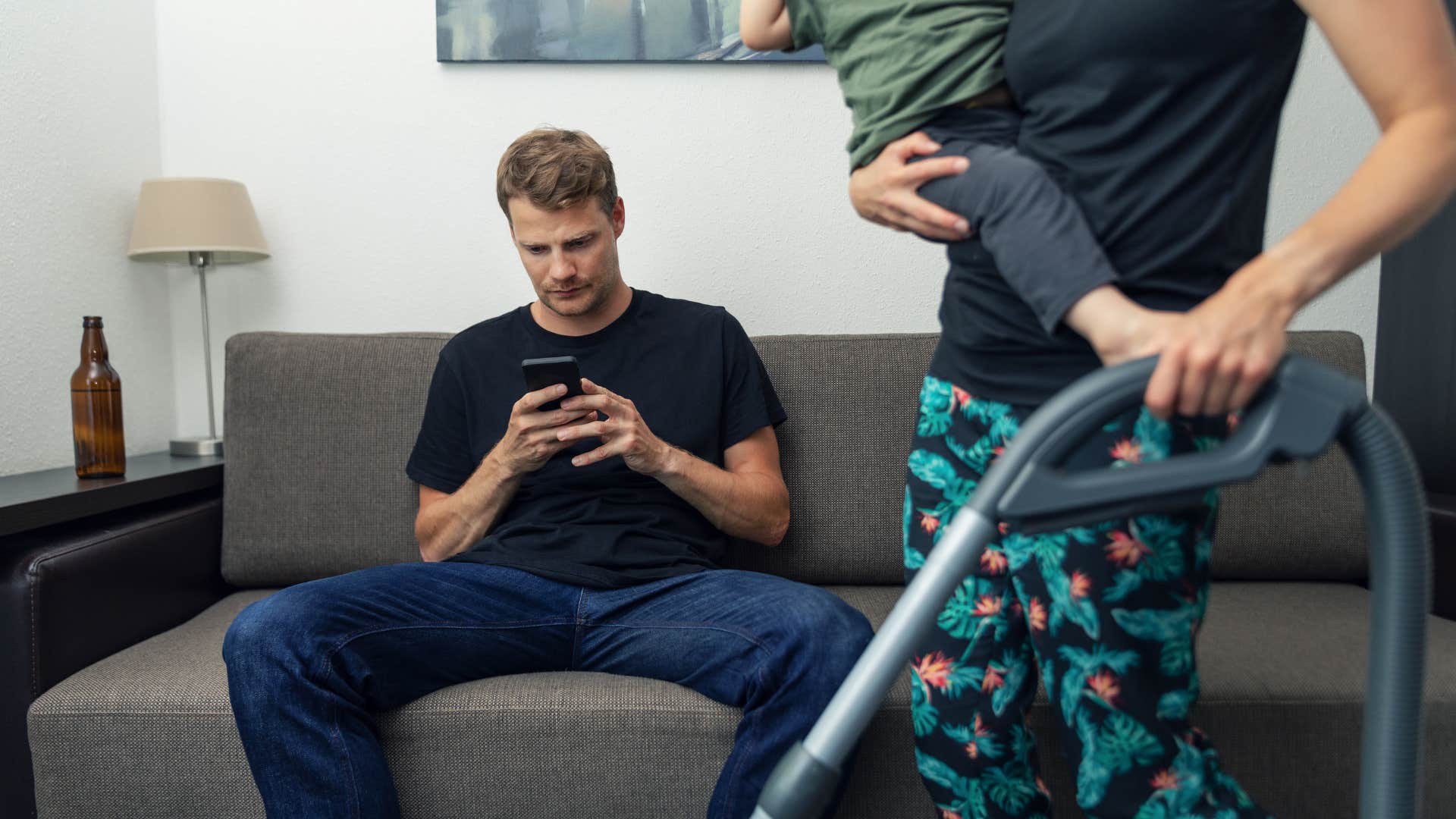 man using weaponized incompetence while wife vacuums holding baby