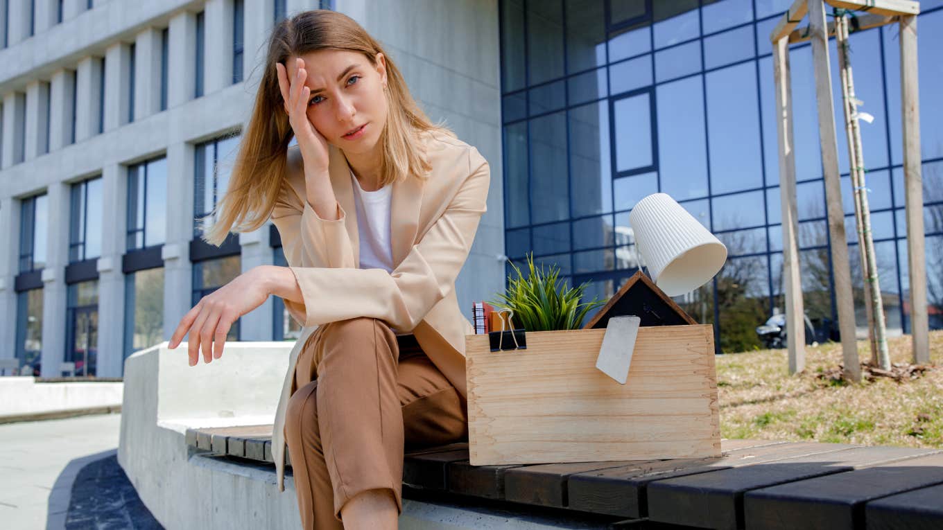 Frustrated woman sitting on bench with box of personal belongings near business center. 