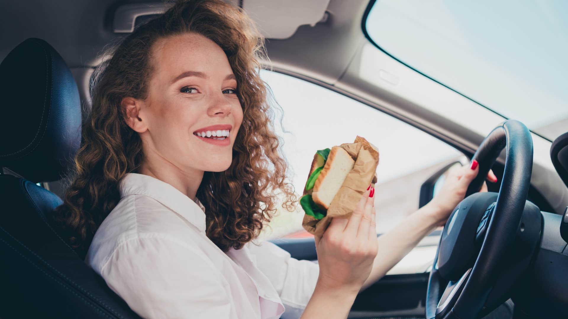 practical woman who eats the same thing for lunch every day driving while holding a sandwich