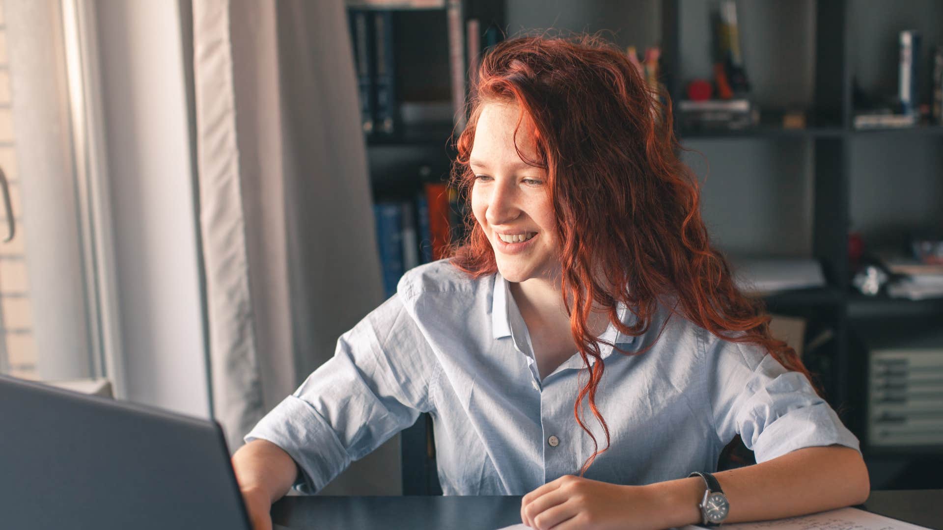 young woman in light shirt writing essays by hand as she looks at laptop